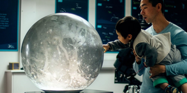Man holds small child up in his arms so that the child can touch the PufferSphere which is displaying the moon. This appears to be happening at a museum.