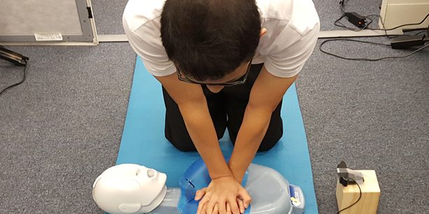 man kneeling on a blue mat performing CPR on a blue and white plastic CPR dummy in front of a webcam